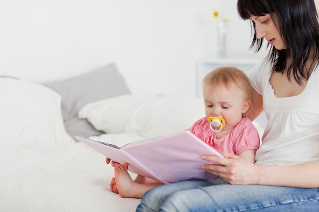 Vrij donkerbruine vrouw die een boek toont aan haar baby terwijl het zitten op een bed