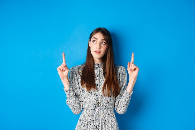 Vrij doordachte vrouw wijzende vingers en denken, peinzende in jurk op blauw.