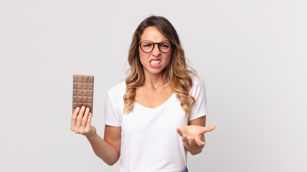 Vrij dunne vrouw die boos, geïrriteerd en gefrustreerd kijkt en een chocoladereep vasthoudt