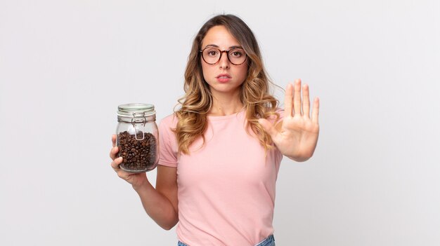 Vrij dunne vrouw die er serieus uitziet met open palm die een stopgebaar maakt en een fles koffiebonen vasthoudt