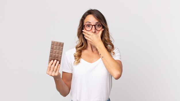 Vrij dunne vrouw die mond bedekt met handen met een geschokte en een chocoladereep vasthoudt