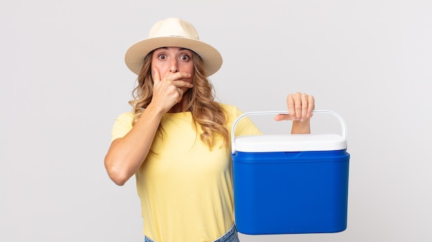 Vrij dunne vrouw die mond bedekt met handen met een geschokte en een zomerpicknickkoelkast vasthoudt