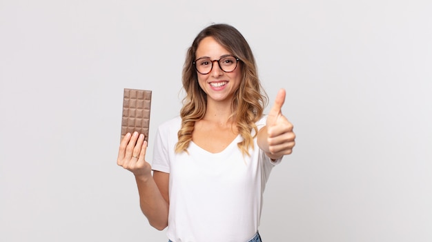 Vrij dunne vrouw die trots is, positief glimlacht met duimen omhoog en een chocoladereep vasthoudt