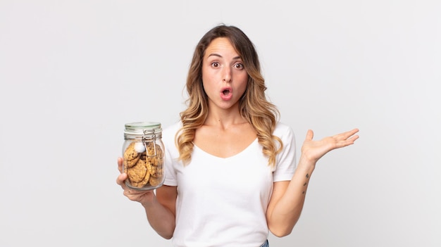 Vrij dunne vrouw die verrast en geschokt kijkt, met open mond terwijl ze een voorwerp vasthoudt en een glazen fles met koekjes vasthoudt