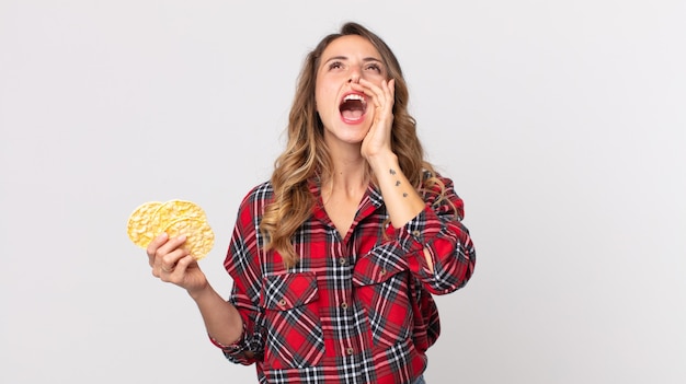 Vrij dunne vrouw die zich gelukkig voelt, een grote schreeuw geeft met de handen naast de mond en een dieetrijstwafels vasthoudt