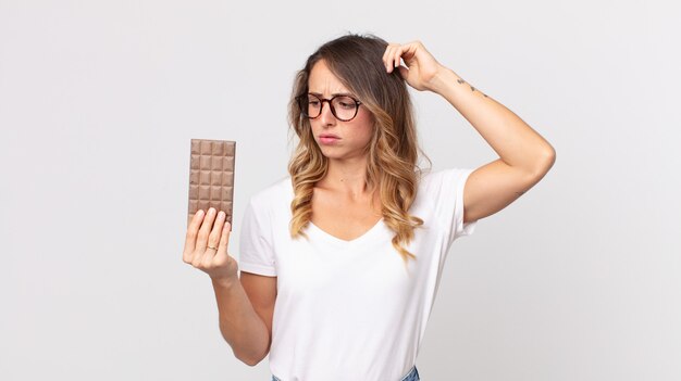 Vrij dunne vrouw die zich verward en verward voelt, haar hoofd krabt en een chocoladereep vasthoudt