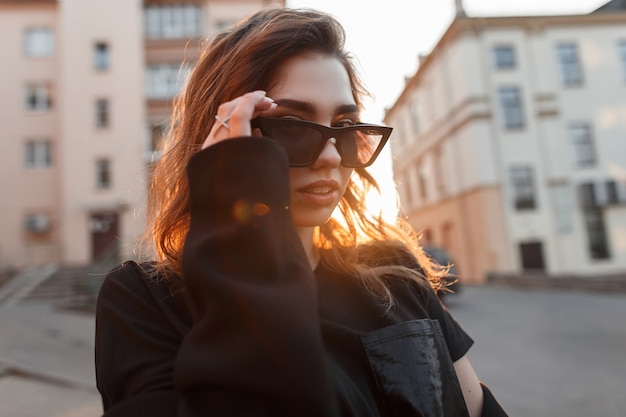 Vrij Europese stijlvolle jonge vrouw hipster in donkere zonnebril in een stijlvolle t-shirt met sexy lippen vormt in de buurt van vintage gebouwen bij zonsondergang