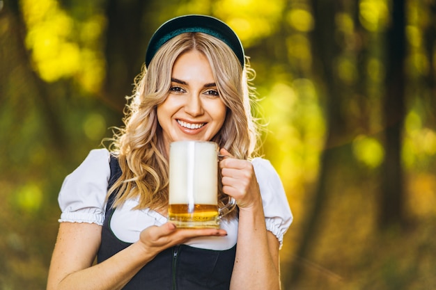 Vrij gelukkige blonde in dirndl, traditionele festivalkleding, die mok bier in openlucht in het bos houden