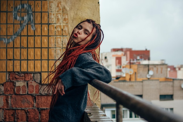 Vrij grappig meisje met rode dreadlocks in een grijze trui die gezichten maakt op het balkon met uitzicht op de stad.