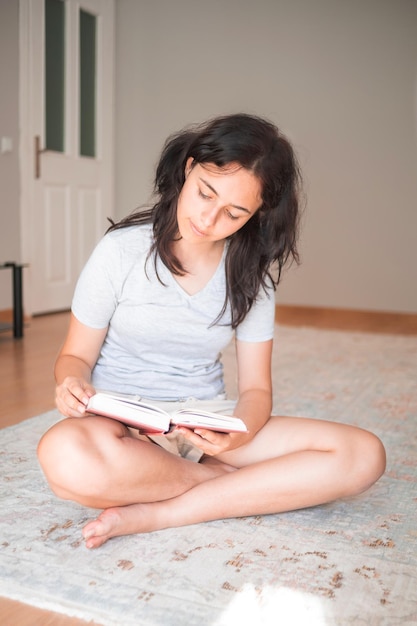 Vrij jong meisje dat een papieren boek leest terwijl ze thuis op een tapijt zit