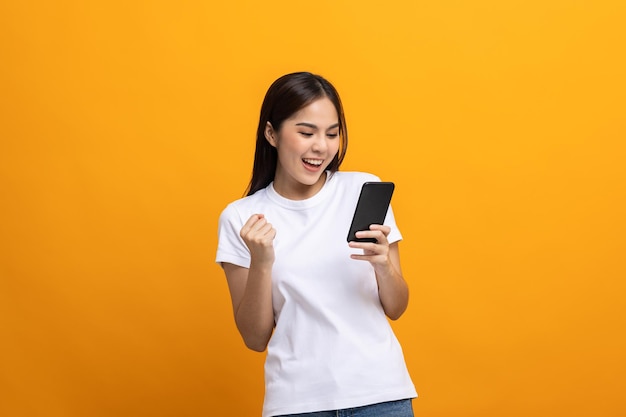 Vrij jonge aziatische vrouw die smartphone gebruikt die zich op geïsoleerde gele achtergrond gelukkig voelt. Winkelen online betaling met mobiele telefoon. Opgewonden en verraste vrouw met mobiele telefoon.