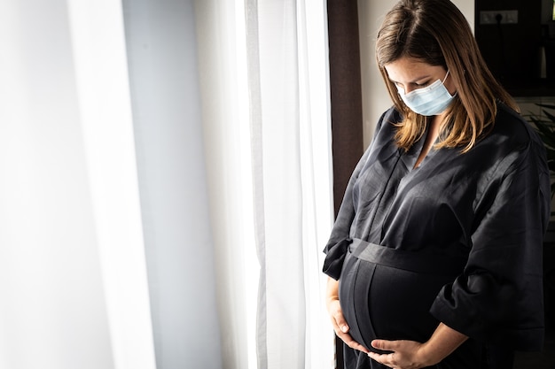 Vrij jonge brunette, zwangere vrouw met een medisch gezichtsmasker tijdens covid-19 pandemie, zwangerschapsconcept