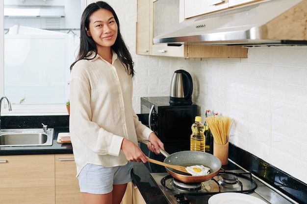 Vrij jonge Chinese vrouw die in pyjama's ontbijt in de keuken kookt