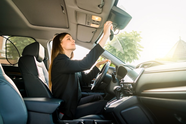 Vrij jonge Kaukasische onderneemster in formele slijtage die in bestuurderszetel zit in haar auto en de centrale achteruitkijkspiegel controleert