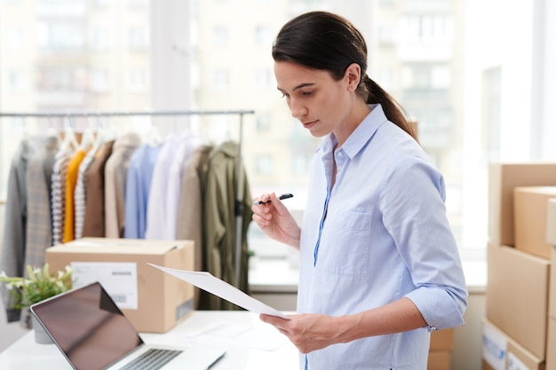 Vrij jonge manager van online winkel die papier met lijst van goederen van één van cliënt leest alvorens de orde in te pakken