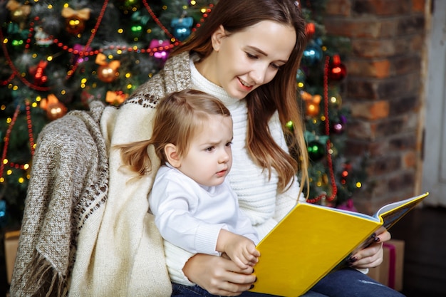 Vrij jonge moeder die een boek binnen leest aan haar leuke dochter dichtbij kerstboom