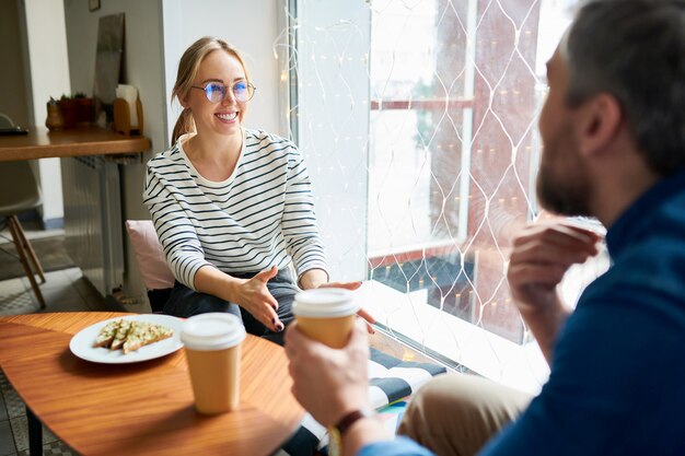 Foto vrij jonge vrolijke onderneemster in vrijetijdskleding en haar collega die koffie hebben tijdens bespreking van werkende vragen in koffie