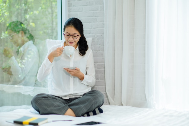 Vrij jonge vrouwenzitting op bed en het genieten van van eerste ochtendkoffie op zonneschijn