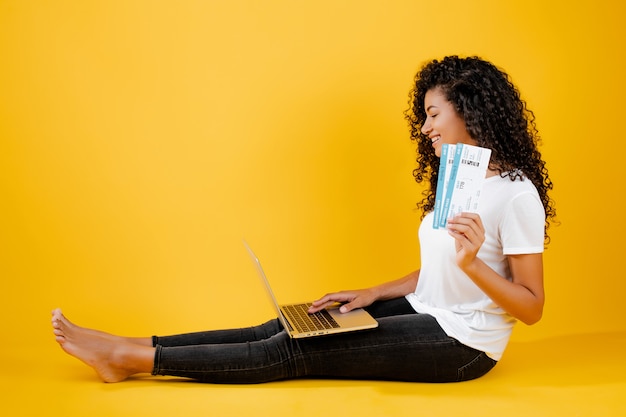 Vrij jonge zwarte Afrikaanse vrouwenzitting met laptop en vliegtuigkaartjes die over geel worden geïsoleerd