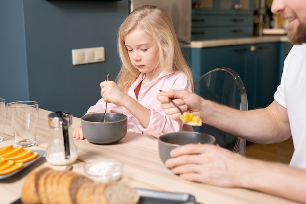 Vrij klein blond meisje en haar vader zitten door houten tafel in de keuken en eten muesli met melk voor het ontbijt in de ochtend