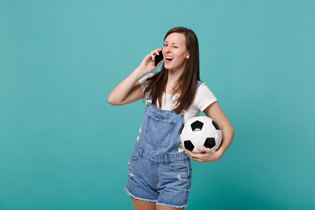 Vrij knipperende jonge meisje voetbalfan met voetbal praten op mobiele telefoon, aangenaam gesprek voeren geïsoleerd op blauwe turkooizen achtergrond. Mensen emoties, sport familie vrije tijd concept.
