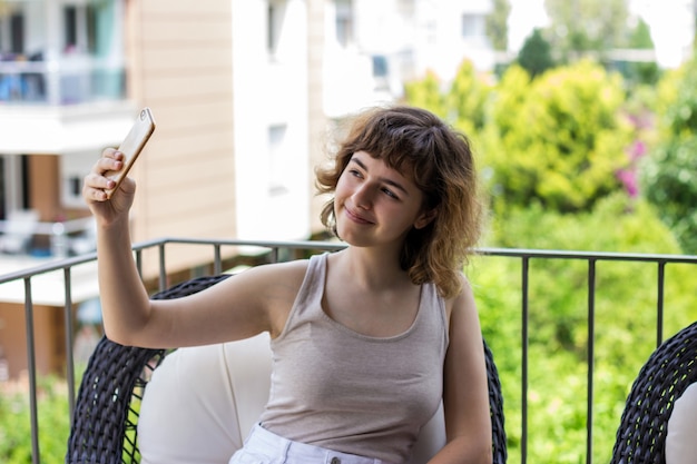 Vrij krullend glimlachend jong meisje dat een selfie neemt op smartphone op het balkon van het appartement. Videochatten met vrienden