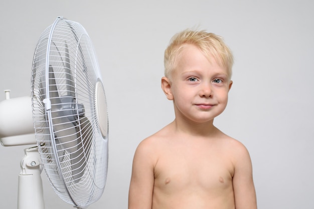 Vrij lachende blonde jongen met naakte torso staat in de buurt van een ventilator. Zomer concept