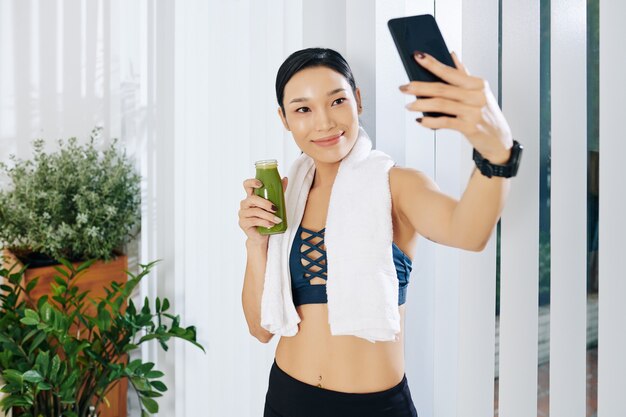 Vrij lachende jonge Aziatische vrouw die groene smoothie drinkt nadat ze 's ochtends thuis heeft getraind en een selfie maakt