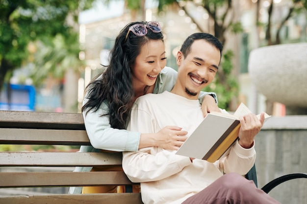 Vrij lachende jonge Aziatische vrouw die in het boek van een vriendje kijkt dat een boek leest wanneer ze op een parkbank zit