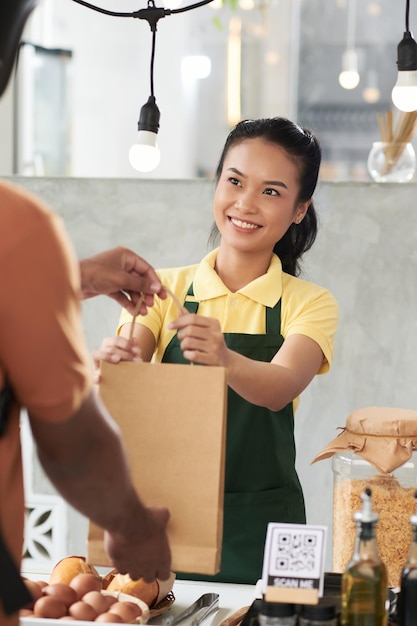 Vrij lachende jonge coffeeshopmedewerker die papieren zak met bestelling geeft aan klant