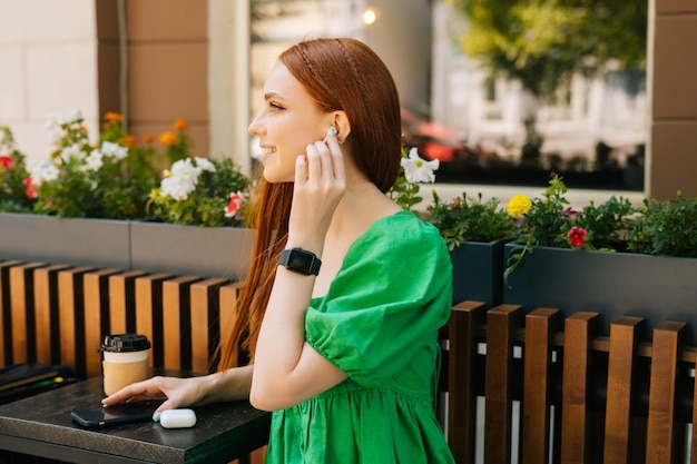 Vrij lachende jonge vrouw die draadloze oortelefoons zet om muziek te luisteren met behulp van mobiele telefoon zittend aan tafel met koffiekopje, op terras in de zomerdag, onscherpe achtergrond, selectieve aandacht