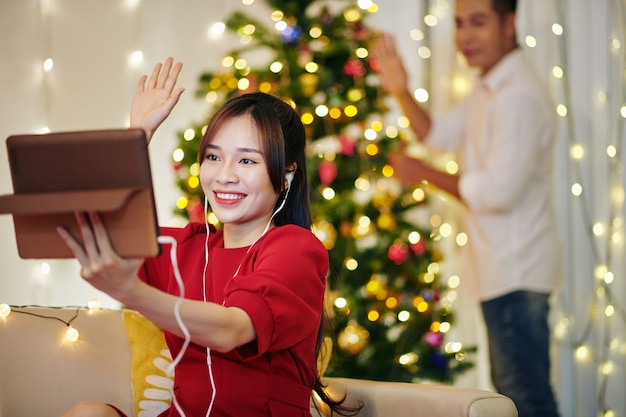 Vrij lachende jonge vrouw en haar vriendje zwaaien met handen wanneer ze hun vrienden videobellen terwijl ze de kerstboom versieren