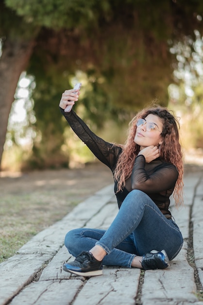 Vrij latijns-vrouw met bril doet een zelfportret met haar smarphone zittend op een houten pad in het veld buitenshuis