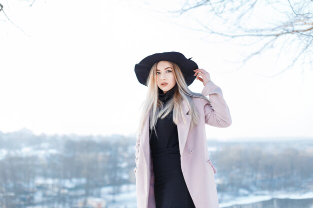 Vrij leuke mooie jonge vrouw met blond haar in een vintage roze jas in een stijlvolle hoed in golf in een rok staat op straat in een zonnige winterdag. Modieus meisje loopt buiten.