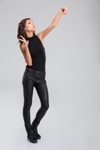 Vrij lieve, krullende jonge vrouw in zwarte leren broek en top die een zelfportret maakt met smartphone en een overwinningsgebaar toont