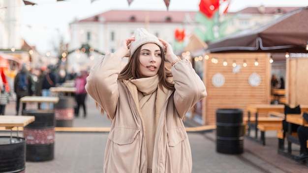 Vrij modieus meisje in stijlvolle winterbovenkleding met een vintage gebreide muts en beige jas met een sjaal lopen in de stad op de beurs