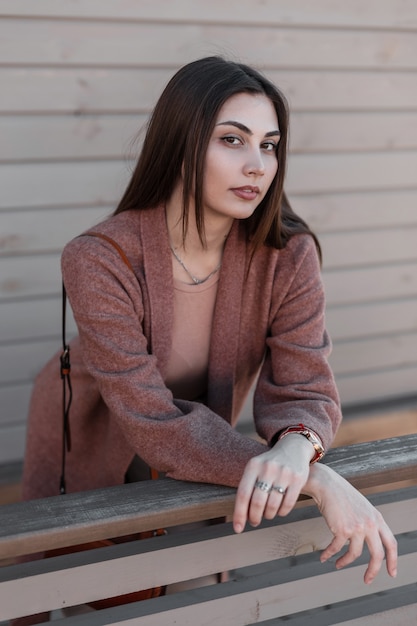 Vrij modieuze aantrekkelijke jonge vrouw met mooie ogen met sexy lippen met bruin haar in stijlvolle jas geniet van vrije tijd staande in de buurt van vintage houten balustrade. Vrouwelijk mooi meisjesmodel buitenshuis.