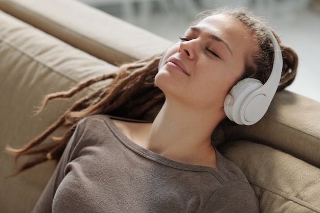 Vrij ontspannen meisje met koptelefoon glimlachen tijdens het luisteren naar rustgevende muziek op de bank na een vermoeiende dag