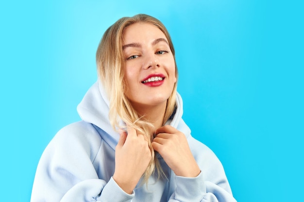 Vrij positief trendy blond meisje in een blauwe hoodie glimlacht en poseert geïsoleerd in modestudio