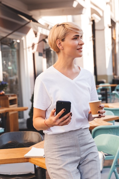 vrij volwassen vrouw, gekleed in wit t-shirt met behulp van mobiele telefoon, zittend in café buiten in de zomer en koffie drinken uit papieren beker