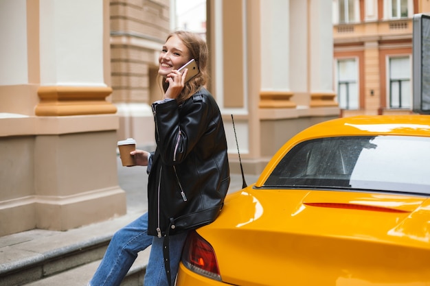 Vrij vrolijk meisje in leren jas met kopje koffie te gaan leunend op gele sport auto praten op mobiel terwijl vreugdevol