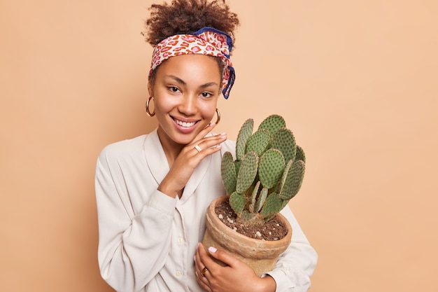 Vrij vrolijke jonge vrouw raakt gezicht zachtjes vast houdt pot sappige groene cactus glimlacht aangenaam toont witte tanden draagt modieuze kleding geïsoleerd over beige muur