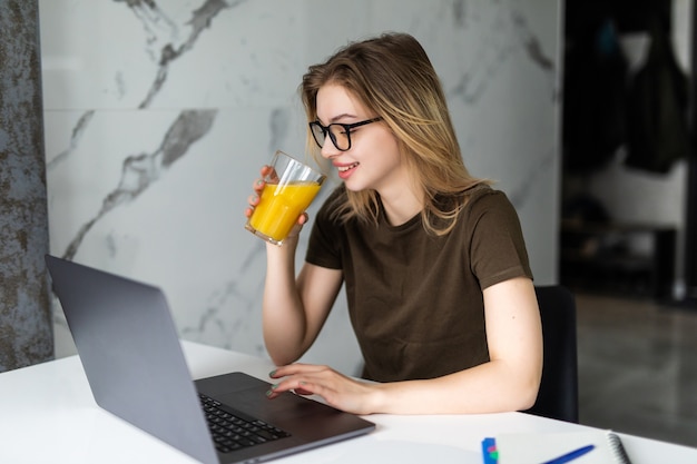 Vrij vrolijke jonge vrouw zittend aan de keukentafel, werken op laptopcomputer, sap drinken