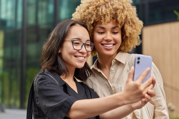 Vrij vrolijke multiraciale vrouwelijke vrienden nemen samen selfie terwijl ze buiten wandelen kijken graag naar de camera aan de voorkant van de mobiele telefoon genieten van vrije tijd gekleed in casual shirts vriendschapsconcept