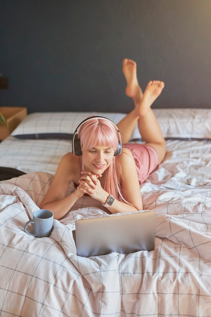 Vrij vrouwelijk model met koptelefoon en beker kijkt naar het scherm van een moderne laptop op bed