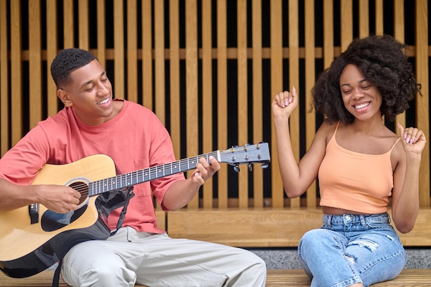 Vrije tijd, gitaar. Vrolijke jonge vrouw met een donkere huidskleur met opgeheven armen en glimlachende man in vrijetijdskleding die gitaar speelt terwijl hij buiten zit op mooie dag