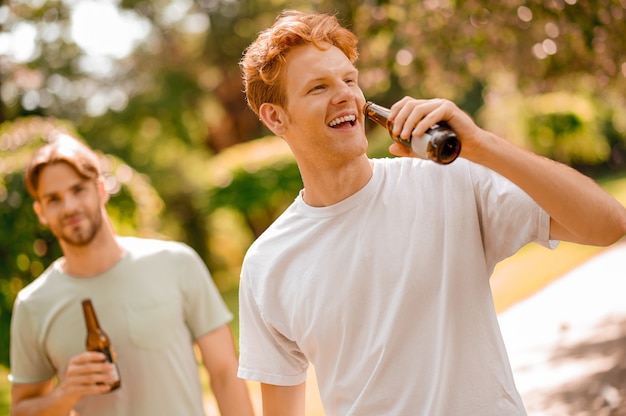 Vrije tijd in de natuur. Vrolijke roodharige man in witte t-shirt met fles drank en vriend achter vrije tijd doorbrengen in de natuur