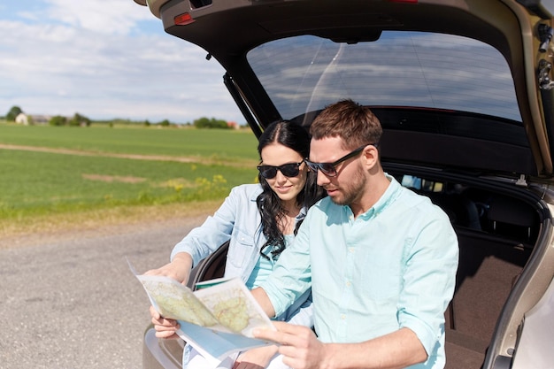 vrije tijd, road trip, reizen en mensen concept - gelukkige man en vrouw zoeken locatie op kaart zittend op kofferbak van hatchback auto buitenshuis