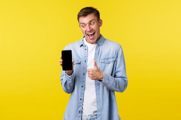 Vrije tijd, technologie en toepassing advertentie concept. gelukkig opgewonden blonde man toont duimen terwijl hij naar het display van de mobiele telefoon kijkt en wijst, terwijl hij nieuwe coole promo of app bekijkt.