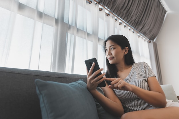 Foto vrije tijd vanuit huis en online winkelconcept, gelukkige aziatische vrouw ontspant en gebruikt mobiele telefoon op de bank in de woonkamer thuis